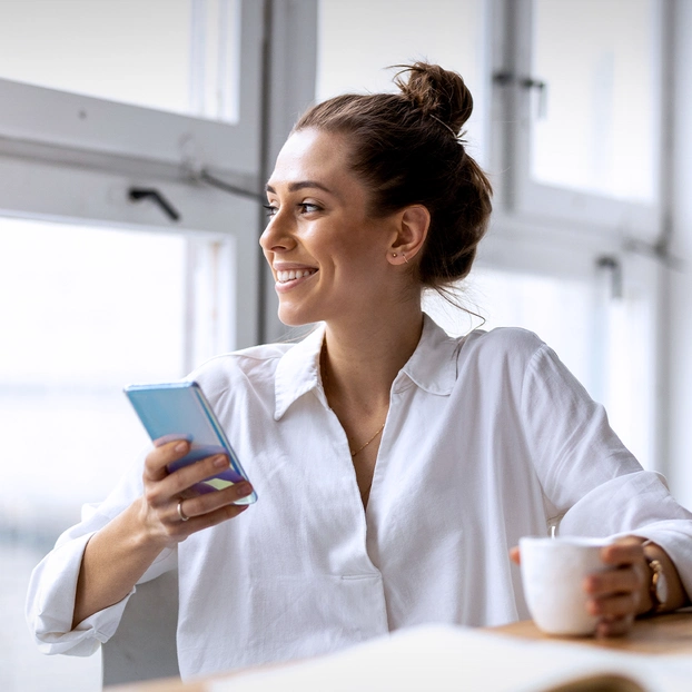 Frau mit Smartphone schaut inspiriert aus dem Fenster