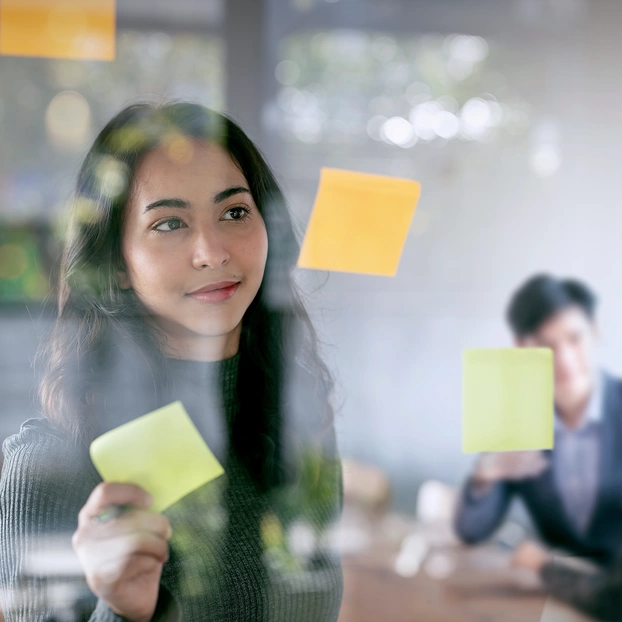 Eine junge Frau bringt in einem Meetingraum Klebezettel an einer Glasscheibe an.