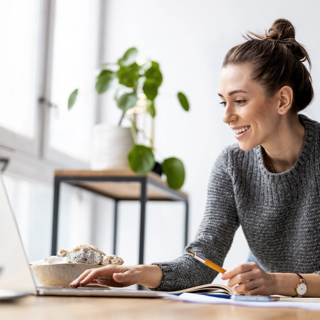 A young woman looks at her laptop. She is sitting in a light-filled room with a cup of coffee next to her on the desk.