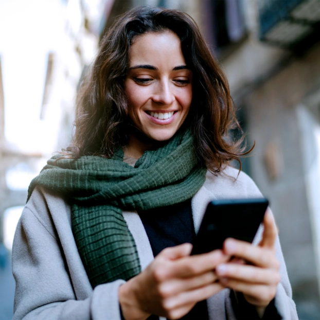 A young woman with a gray coat and a green winter scarf in a narrow street. She looks at her smartphone with a smile.