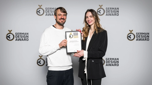 Nicole Lössner und Philipp Hahn von ]init[ mit der Urkunde des German Design Award vor einer Fotowand bei der Preisverleihung.
