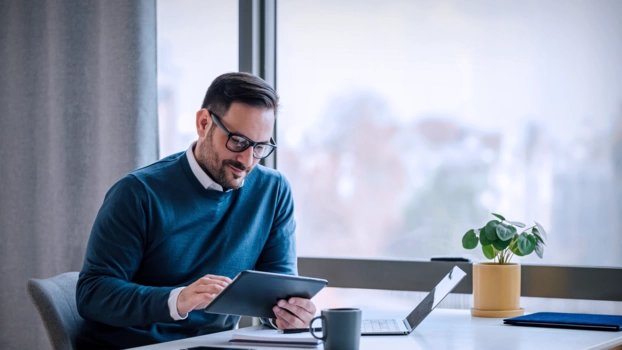 A man sits at a desk and uses the tablet for research. In the background you can see a large window with a view of a city.